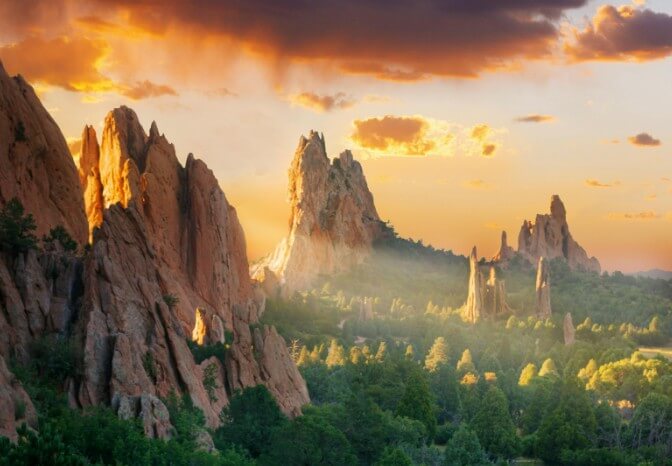 View of Garden of the Gods park in Colorado Springs at sunset. Large red sandstone rock formations rise from a forested area. The sky contains scattered clouds lit by the setting sun, which creates illuminated shafts through the rock formations. The vegetation in the foreground consists of pine trees and other native plants.