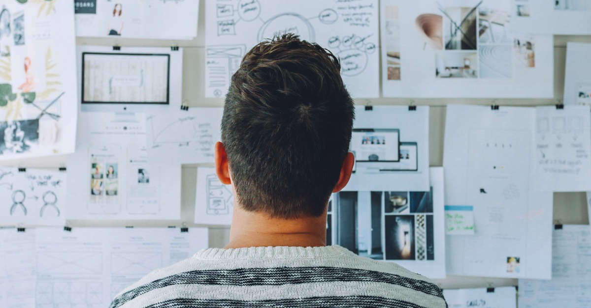 A person stands facing a wall covered with papers and diagrams, appearing to analyze or study the content.